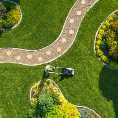 Person mäht Rasen in einer gepflegten Gartenlandschaft mit Rundwegen.
