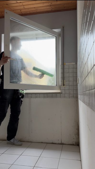 Person reinigt ein Fenster in einem hellen Raum mit Fliesenwand.